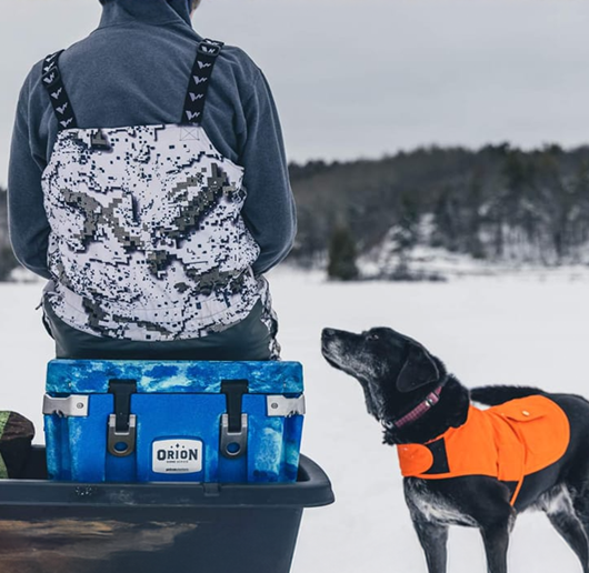 a man sits on his Orion Cooler with his black bird dog at his side