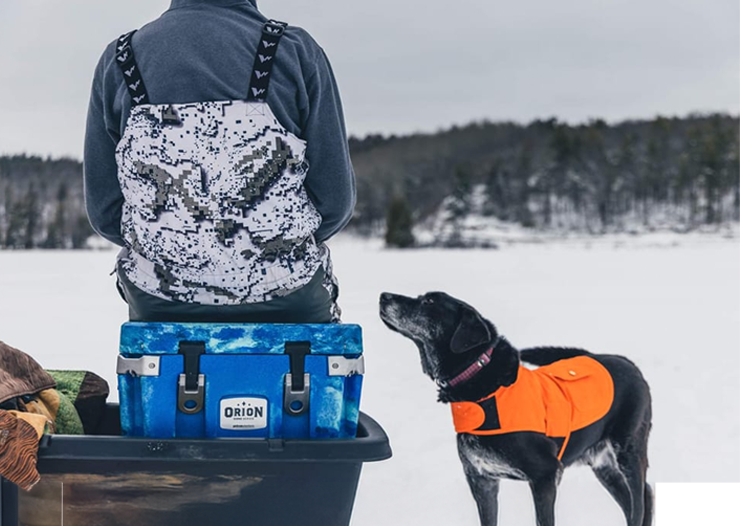 a man sits on his Orion Cooler with his black bird dog at his side
